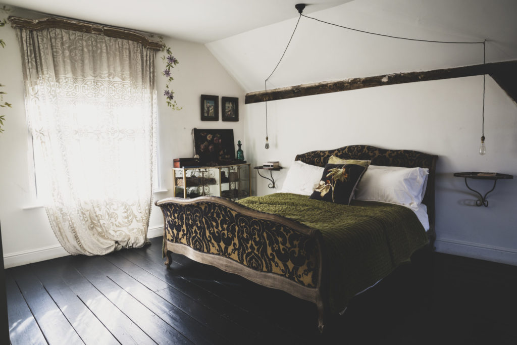Interior view of attic bedroom with dark wooden floor and white walls, antique French double bed with green throw and lace net curtain in window.